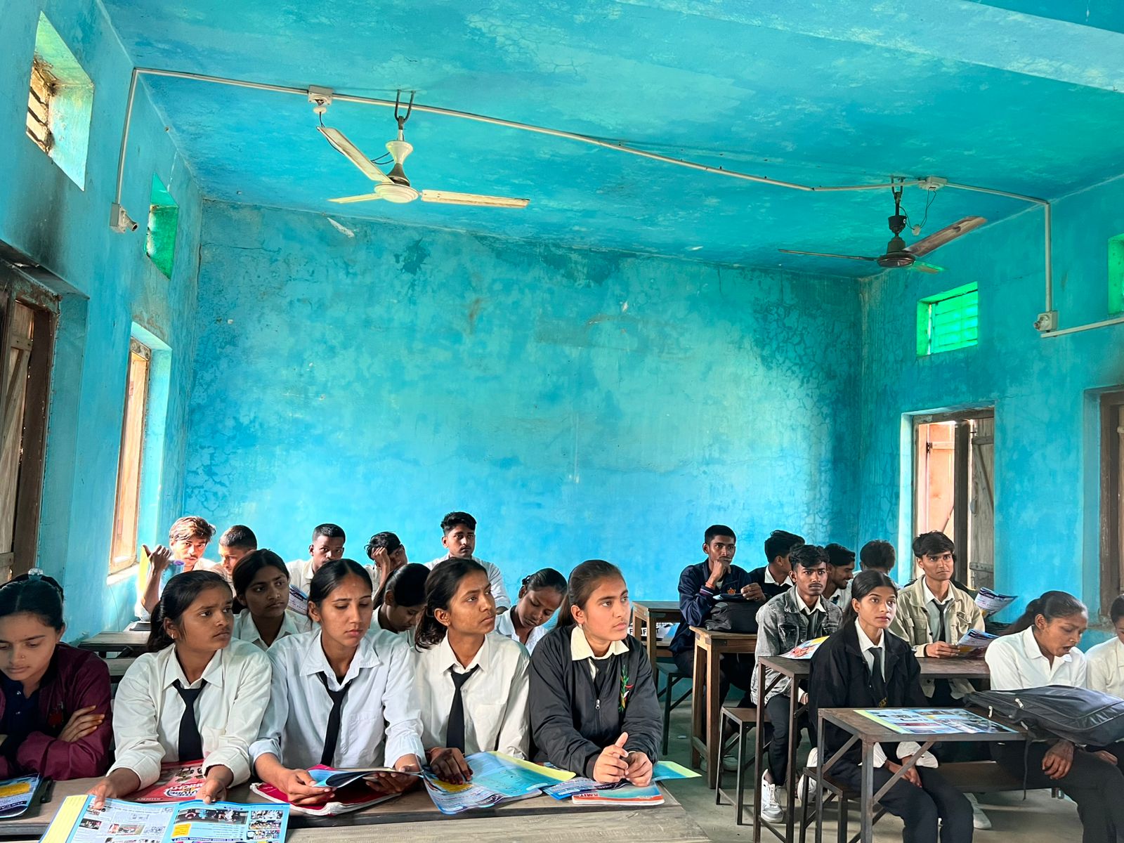 Students sitting attentively in a blue classroom holding Mission Prayas materials